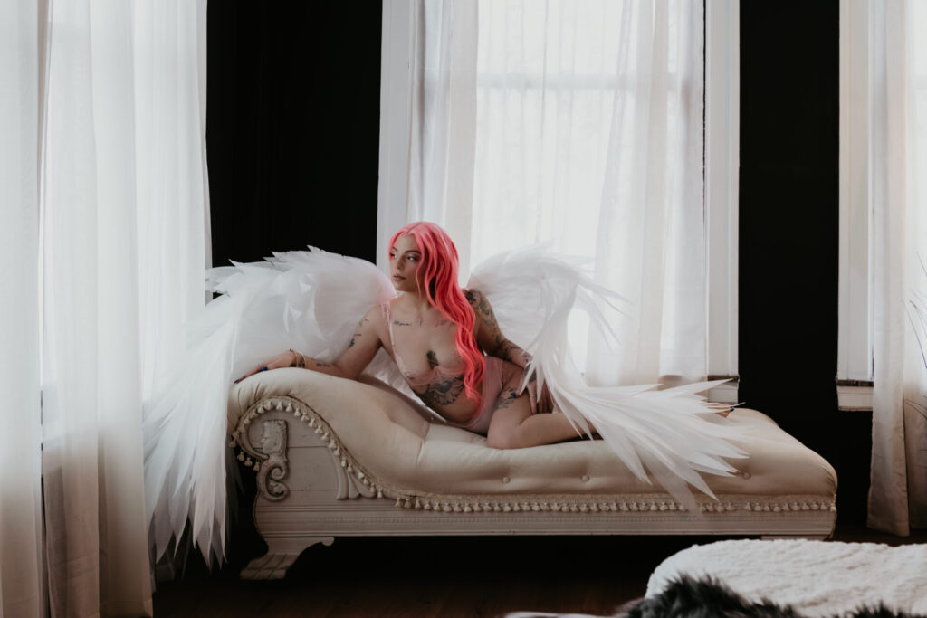 Woman with pink hair in blush lingerie and angel wings posing on a white chaise lounge in a boudoir studio