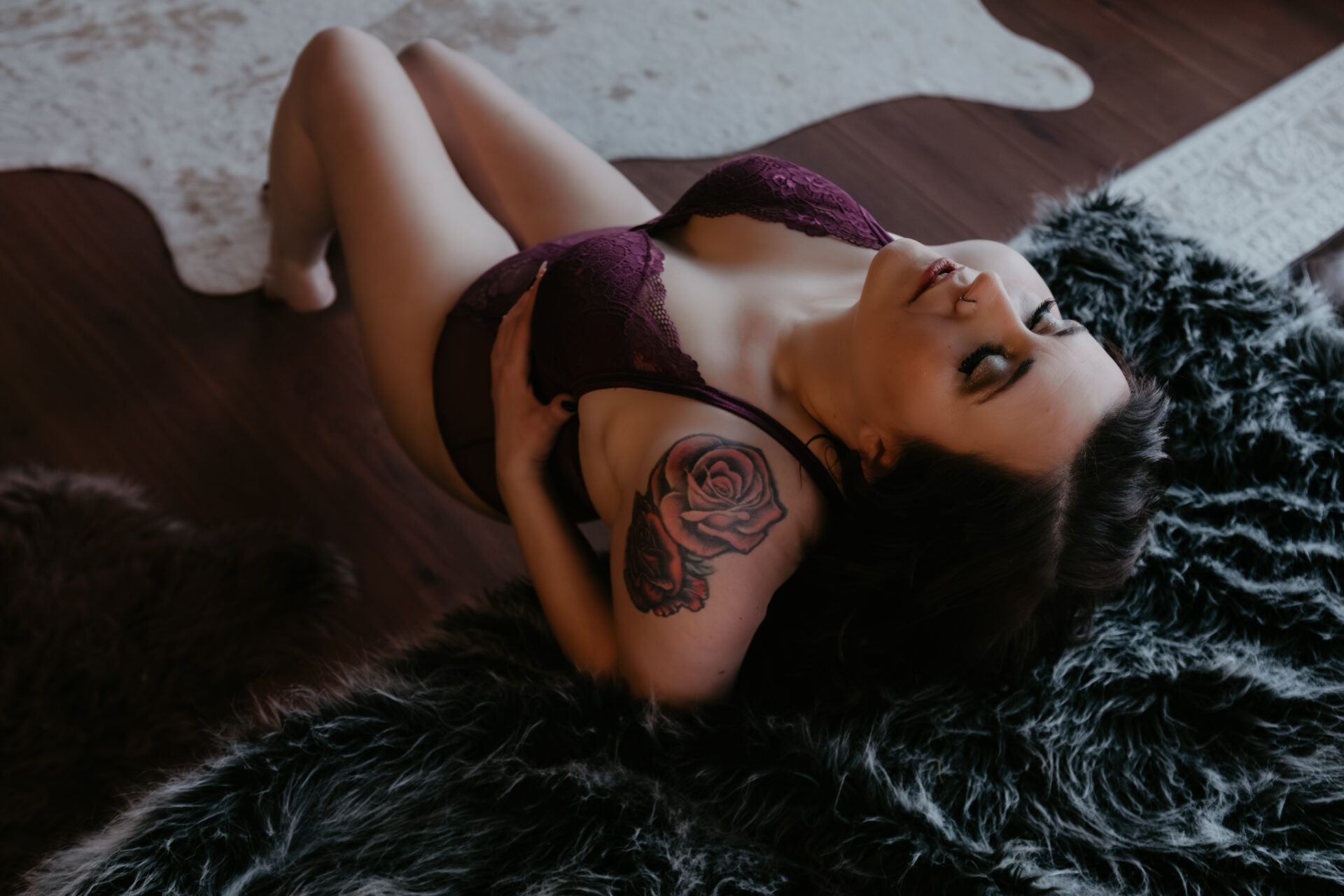 Boudoir photo of a woman reclining on a fur rug wearing burgundy lace lingerie with eyes closed in a Bay City Michigan studio, client traveled from Clare Michigan