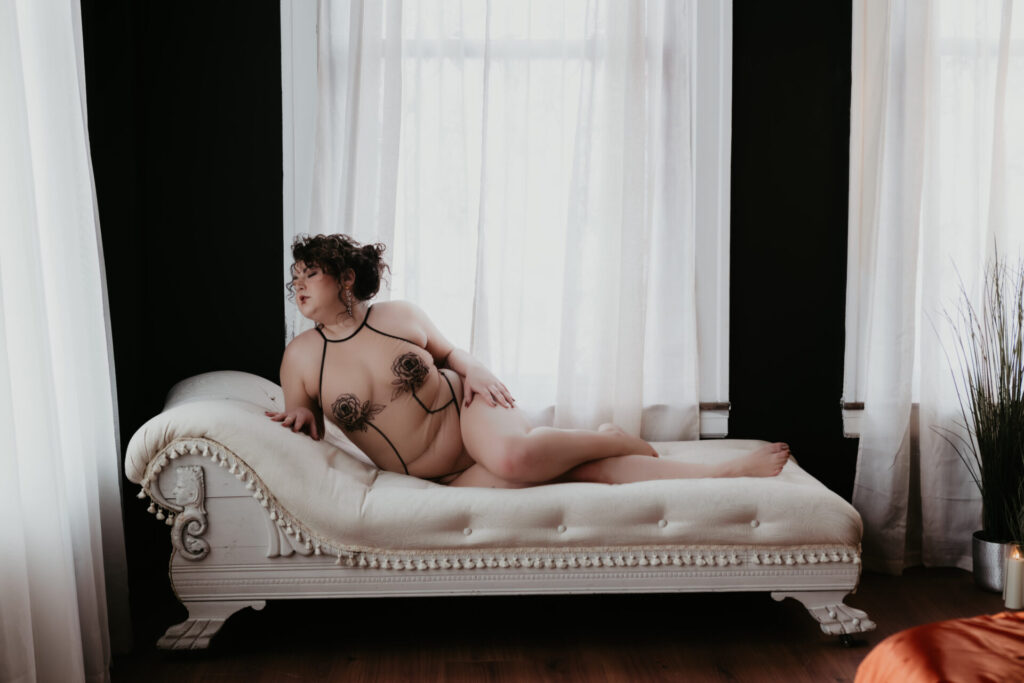 Curvy boudoir client posing on a white vintage chaise lounge in sheer floral lingerie with soft natural window light in a luxury Michigan boudoir studio.