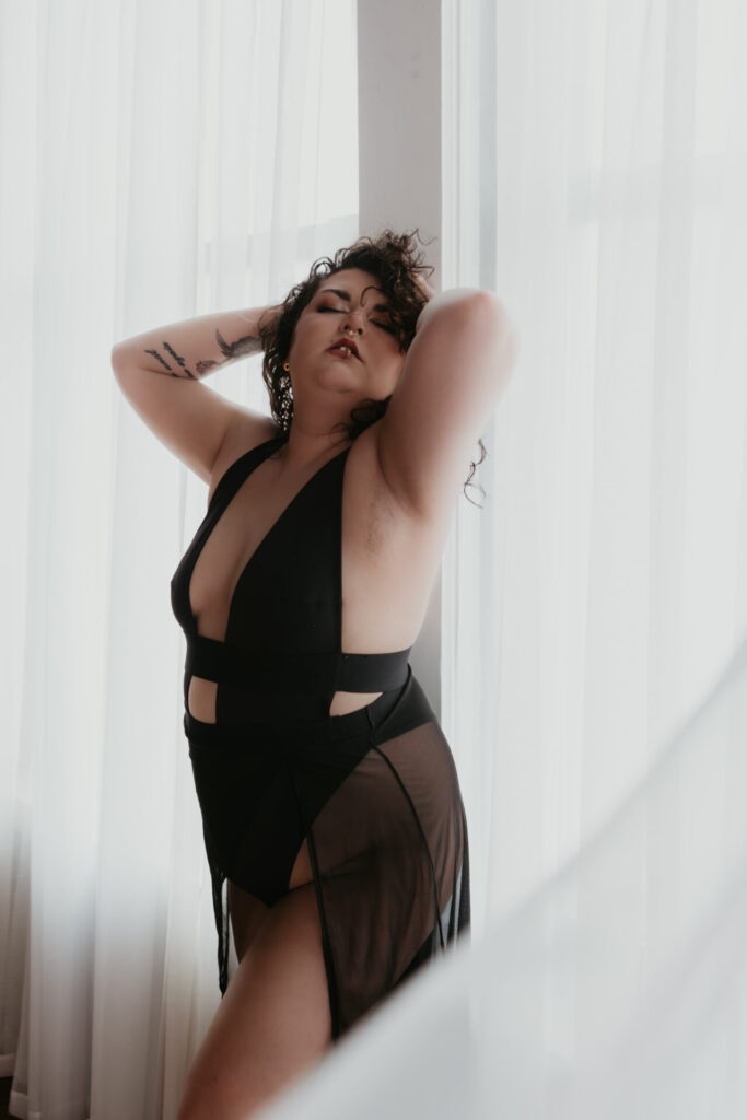 Curvy boudoir client posing in black sheer lingerie between white flowing curtains with soft natural light in a Bay City Michigan boudoir studio.