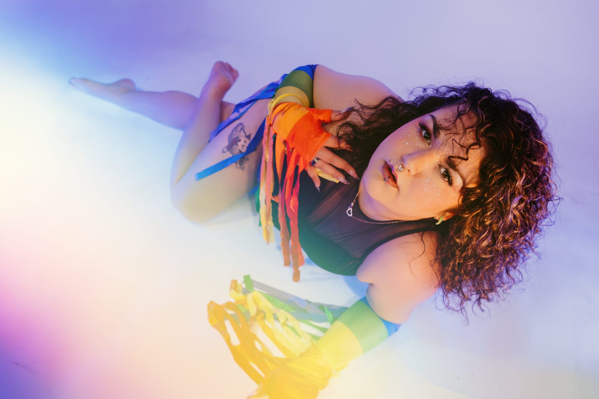 LGBTQ+ pride boudoir portrait featuring a femme client in rainbow fringe gloves and matching lingerie posing on a white floor with colorful lighting during Pride boudoir mini sessions at Nice & Naughty in Saginaw, Michigan by Arielle Roth Boudoir.