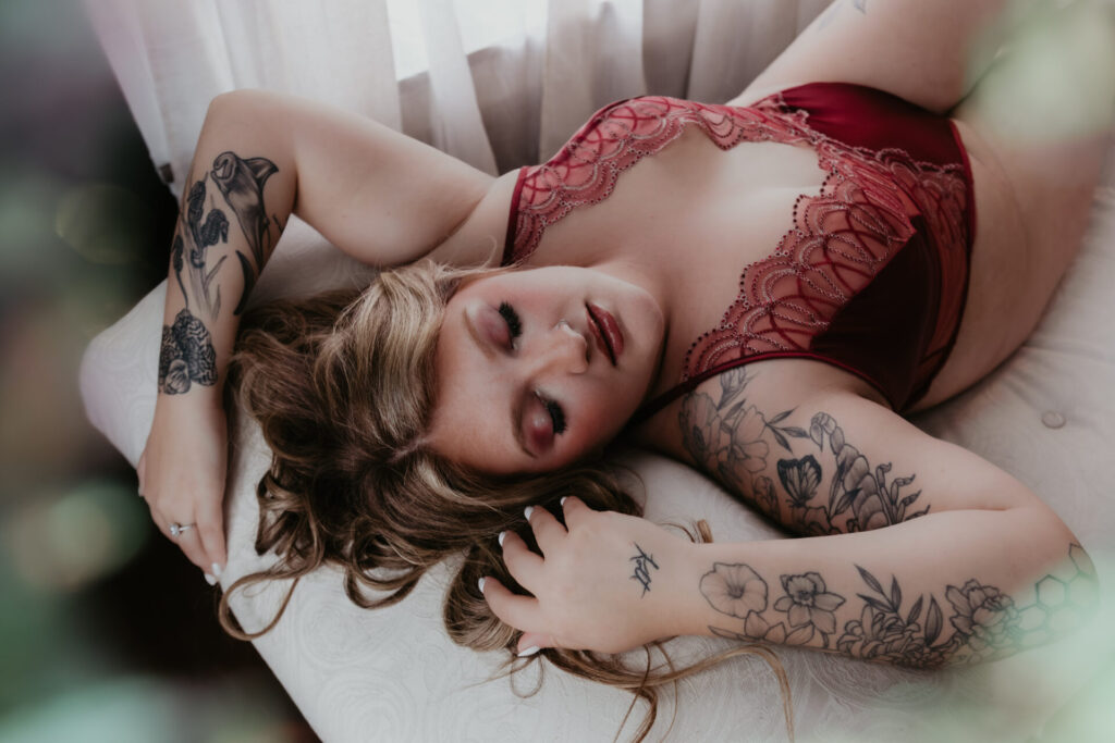 Plus size boudoir client in red lace lingerie laying across a chaise lounge during a romantic boudoir session in Bay City, Michigan.